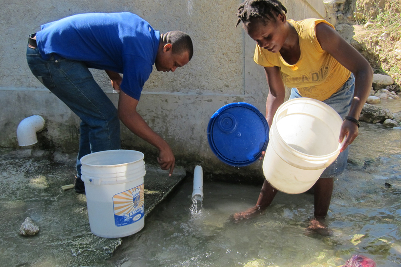 A community member gathers water from a spring in the village of Trianon, Haiti, as a member of DINEPA (the National Directorate for Drinking Water and Sanitation) tests the water.