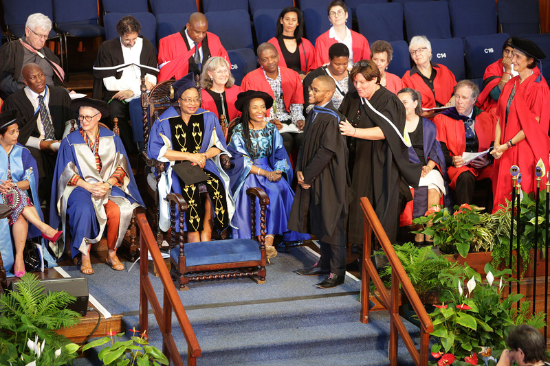 Gerda Kruger, Executive Director: Communication and Marketing Department, hoods her son Siya Kruger at the Humanities graduation event, as Chancellor Graça Machel and VC Prof Mamokgethi Phakeng look on.