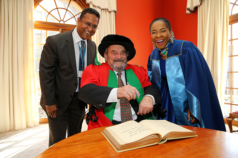 Honorary doctorate recipient Denis Goldberg and VC Prof Mamokgethi Phakeng share a light moment with Registrar Royston Pillay during the signing of the book of honour.
